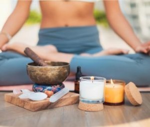Parte de baixo de uma mulher branca, da barriga para baixo, aparece sentada com as pernas cruzadas em pose de yôga, com cuba tibetana em sua frente e algumas velas aromáticas e cristais.