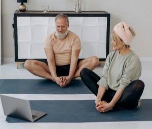 Um homem de barbas branca e camisa creme e bermuda escura, e uma mulher de cabelos grisalhos também, camiseta clara esverdeada, e calça azul marinho, ambos sentados em posição borbleta e segurando as pontas dos pés como se estivessem alongando assistindo a algo no computador notebook que estão no chão a frente deles.