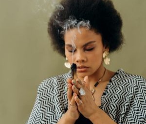 Mulher negra, corte de cabelo afro, olhos fechados em pose meditativa, com mãos sobrepostas segurando um bastão de incenso queimando e soltando fumaça.