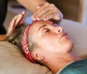 Mulher branca deitada com faixa vermelha prendendo os cabelos na cabeça, olhos fechados, loira, recebendo reiki através da imposição de mãos, uma mão sobre sua fronte, e outra no topo da cabeça, sem encostar.